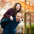 Couple photography in a park with the man giving the woman a piggyback ride, both smiling against a blurred autumn background.