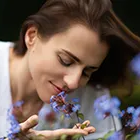 Close-up portraiture of a woman gently smelling a small bunch of purple/blue wildflowers outdoors.