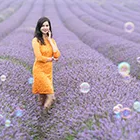 Mid-torso portrait of a woman in a bright orange dress standing in a lavender field with colorful bubbles floating around her.