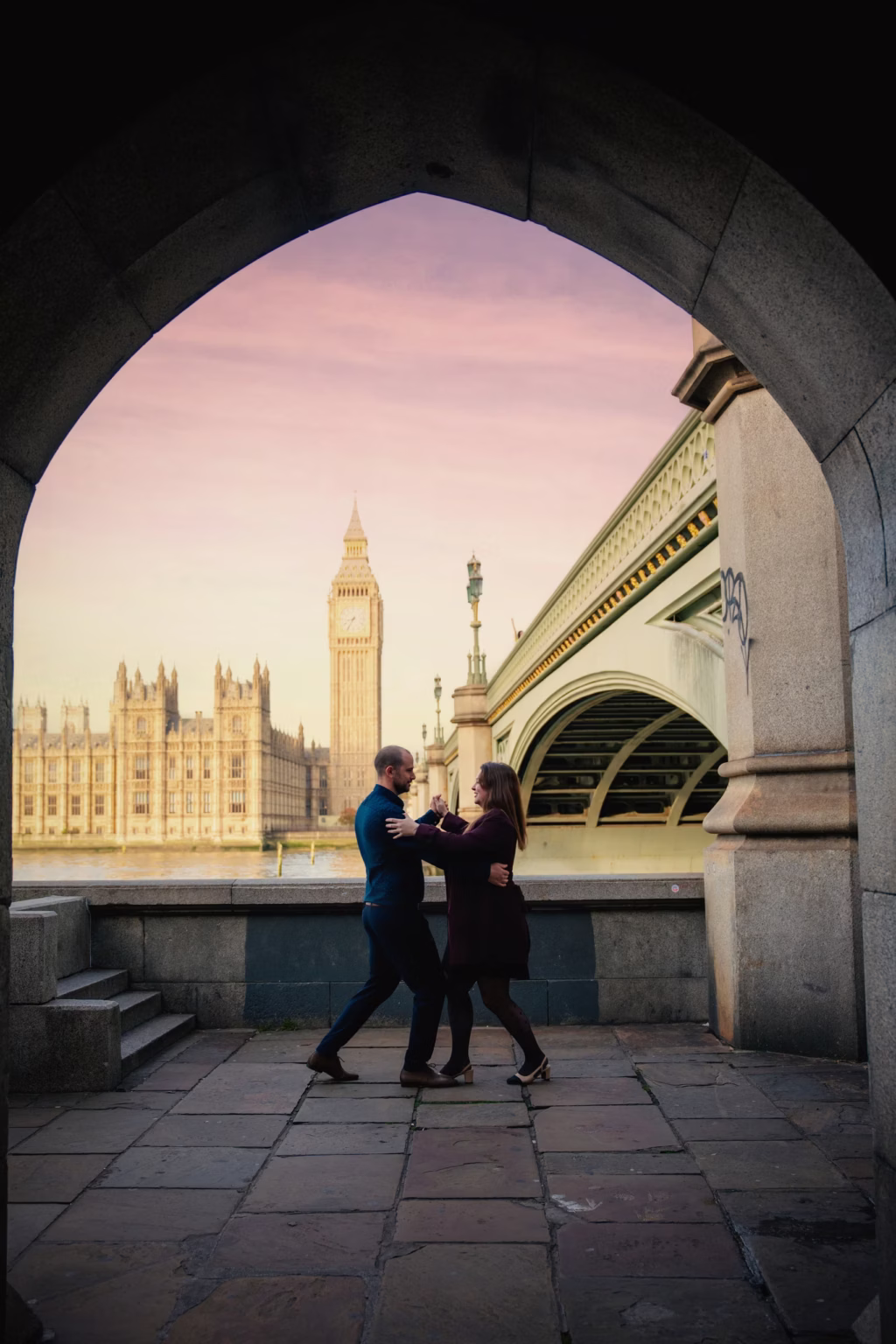 couple london scenic shot framed through Gothic stone archway with couple dancing on paved embankment, Westminster Bridge and Big Ben in background against soft painted purple and orange sunset