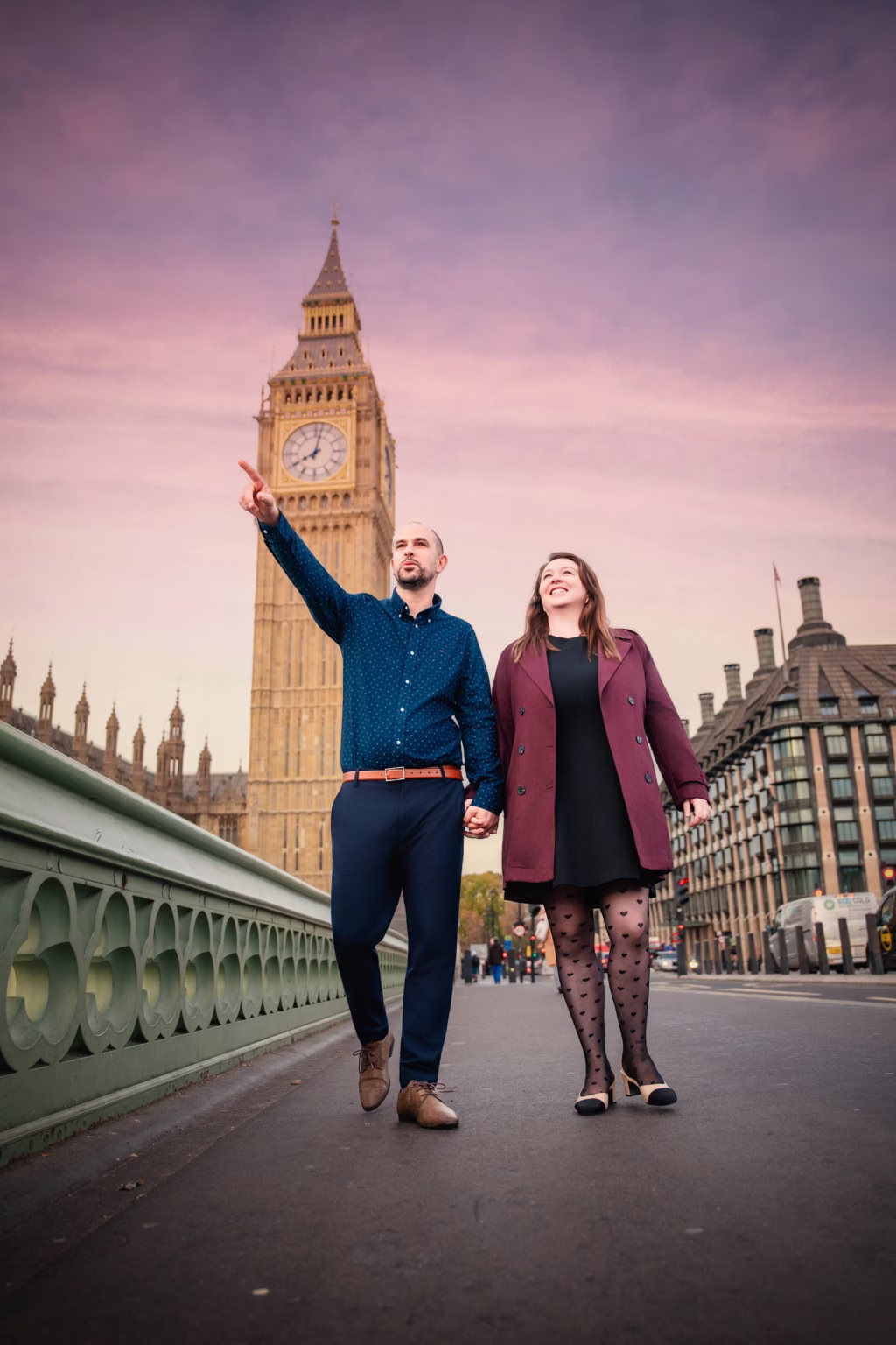 Of london low-angle shot of couple walking along bridge, man in blue patterned shirt pointing toward horizon with woman in plum coat smiling, Big Ben towering behind under vivid pink and purple sky - beautiful couple photoshoot