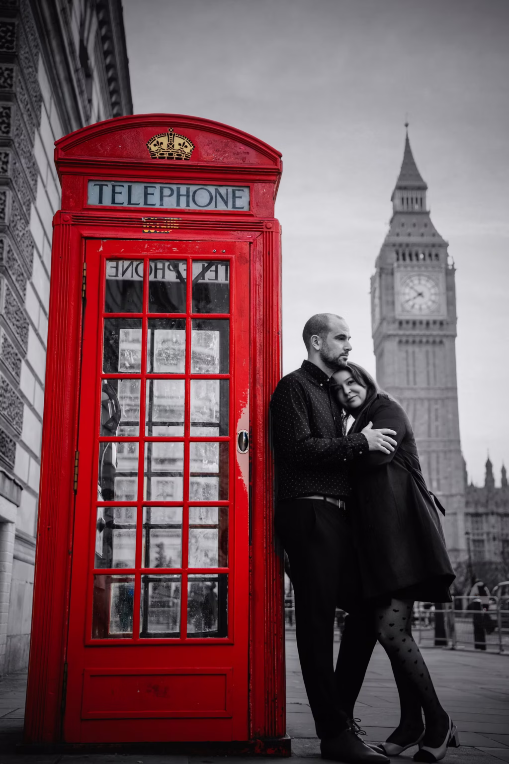 Black and white stylized photo of couple leaning against bright red London telephone box, red booth and gold crown only colors in selective color image, Big Ben visible in blurred background - couple photographers
