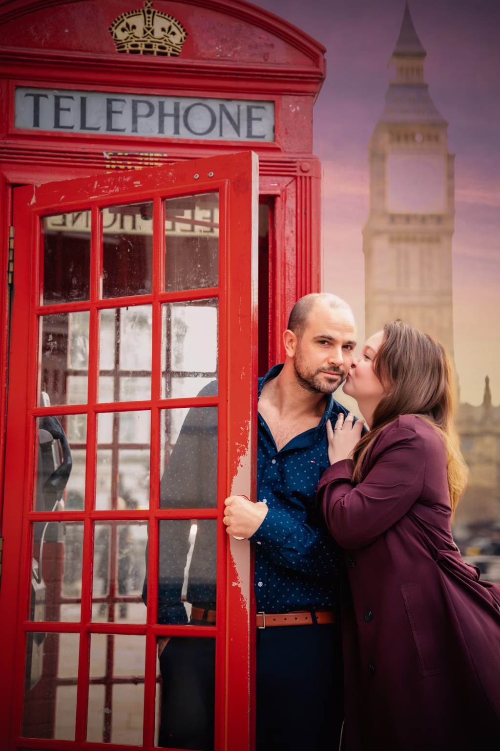 Warm sun drenched shot of couple at red telephone booth, man standing inside open door with woman leaning in to give him kiss on cheek, blurred golden hued Big Ben in background - couple photoshoot photographer