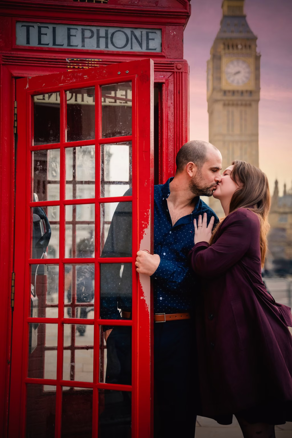 couples photoshoot london romantic close up of couple sharing kiss by red telephone box, man framed within door of booth with woman leaning toward him, soft focus view of Big Ben under twilight sky in background