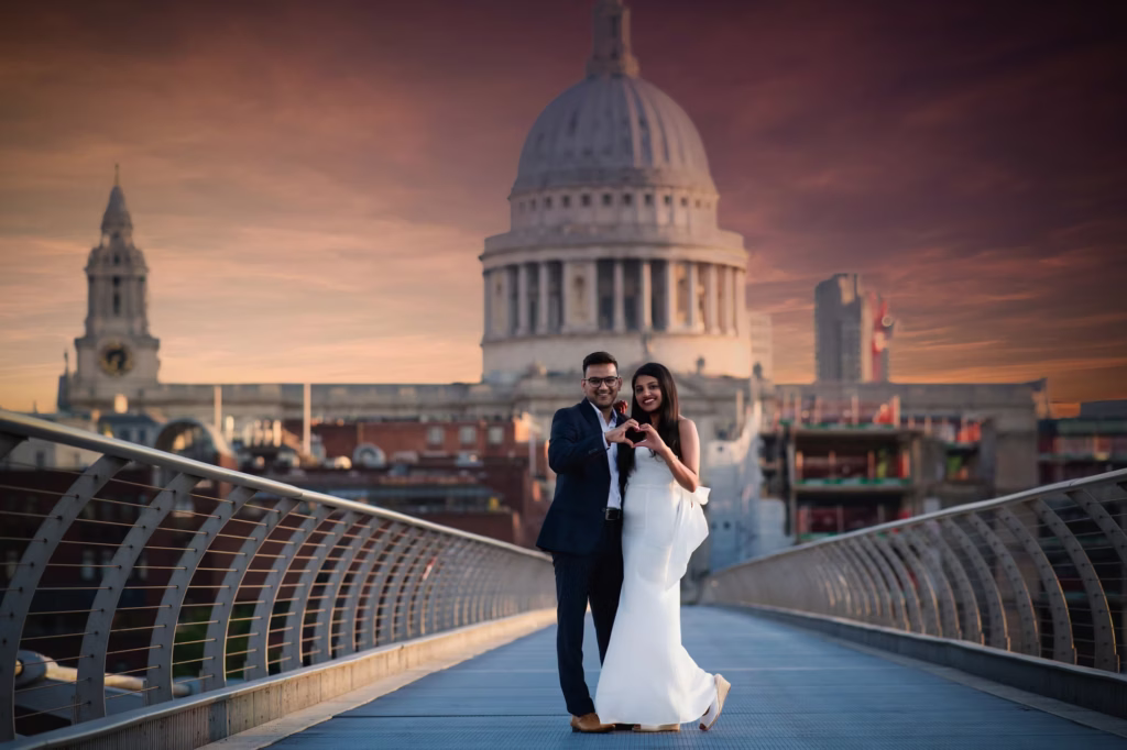 Medium shot of couple on Millennium Bridge facing camera forming heart shape with hands against large St. Paul's Cathedral dome under vivid twilight sky - best engagement photos