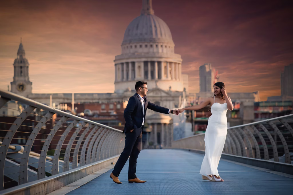 Candid moment on Millennium Bridge with man leading woman by hand looking back at her with smile, St. Paul's Cathedral and Northbank skyline visible under deep red and orange sunset - engagement shoot