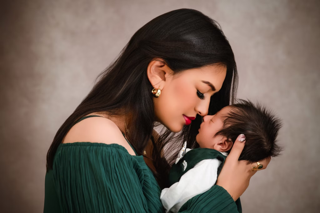 Profile studio shot of mother in dark green dress touching nose to nose of sleeping newborn, baby with dark spiky hair swaddled in green, mottled warm grey background - family photographer london profile green nose touch