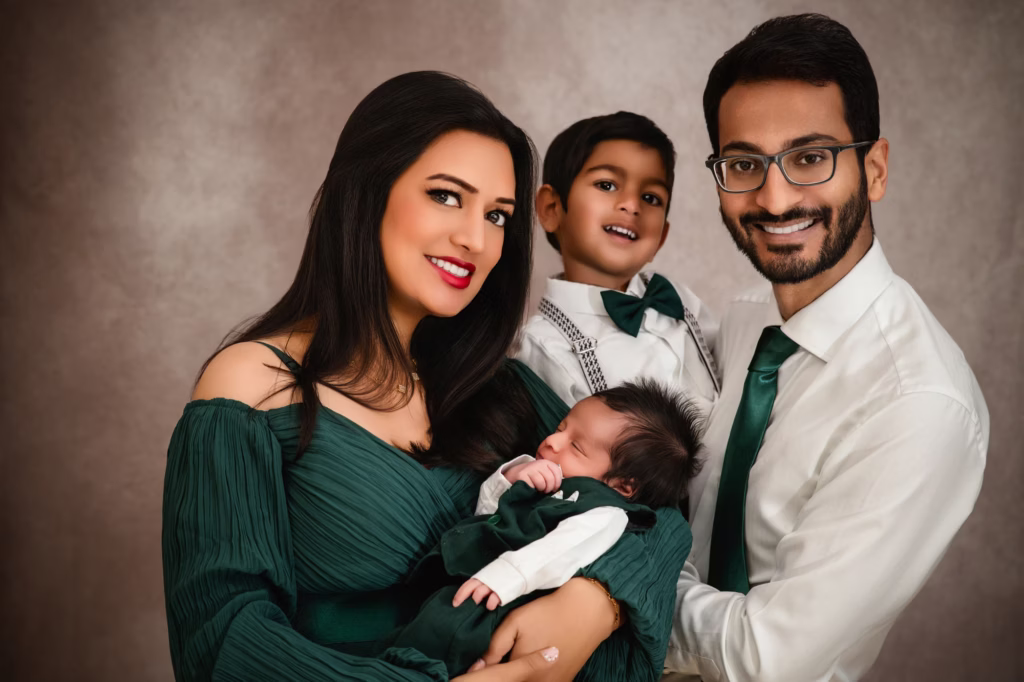 Full family studio portrait against textured beige background, parents and older son smiling while mother holds sleeping newborn, family color-coordinated in forest green and white - family photographer london, coordinated