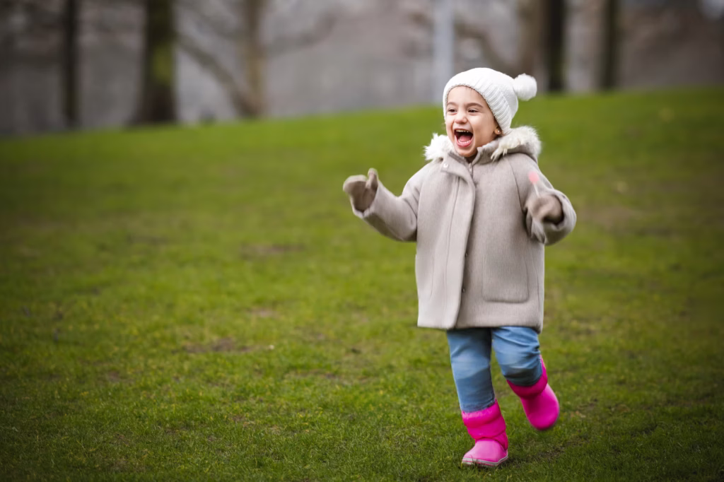 Young child in beige winter coat blue jeans bright pink boots white pom-pom hat running through grassy park with ecstatic open-mouth laugh expression and blurred trees background - family photographer