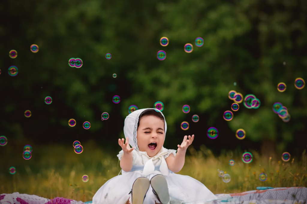 Baby girl in white dress and bonnet surrounded by iridescent bubbles in field with open-mouthed delighted expression hands raised - family portrait photoshoot