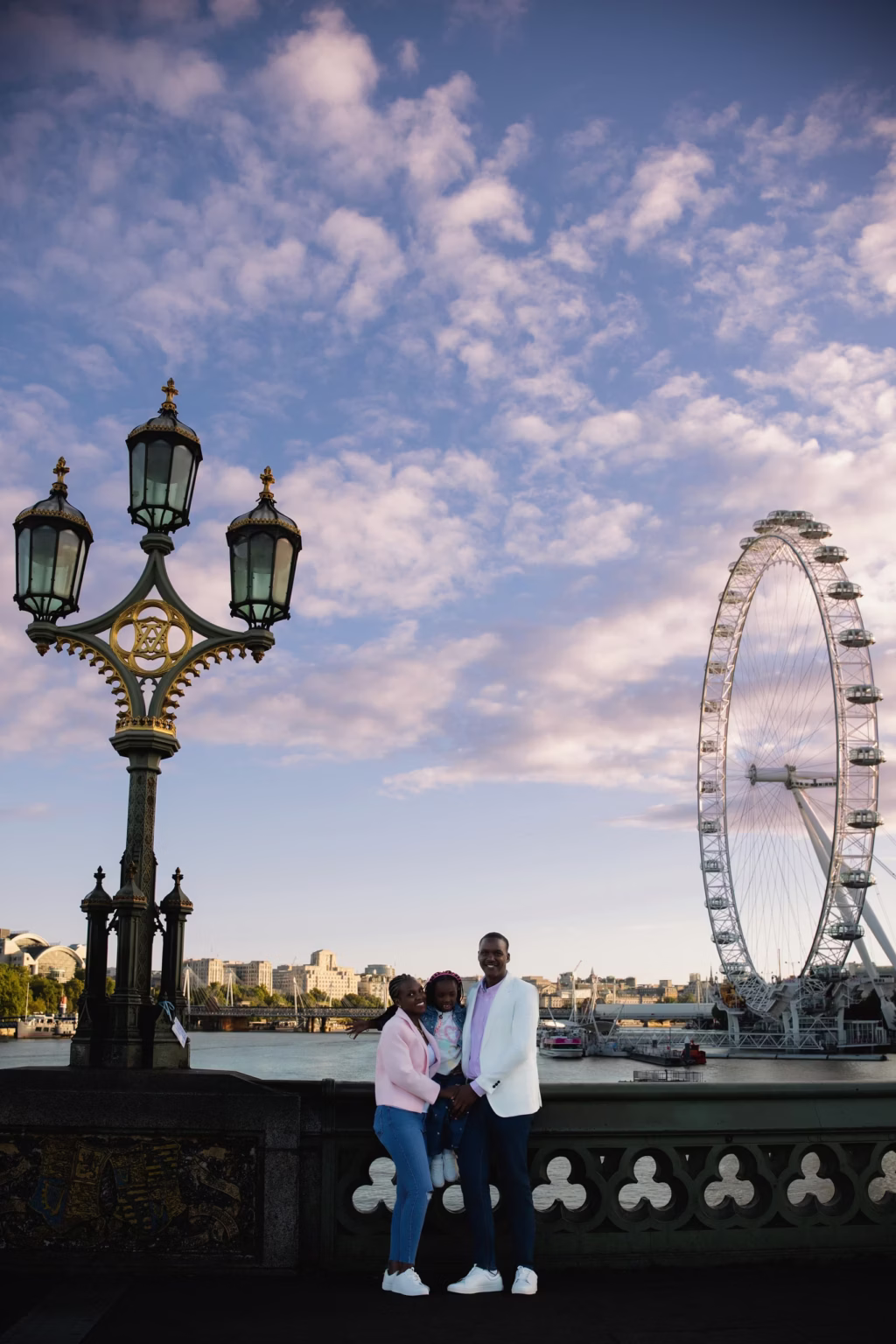 Of three mother father and young daughter standing together on Westminster Bridge with London Eye prominently featured in background under blue sky with soft clouds - family portrait photoshoot