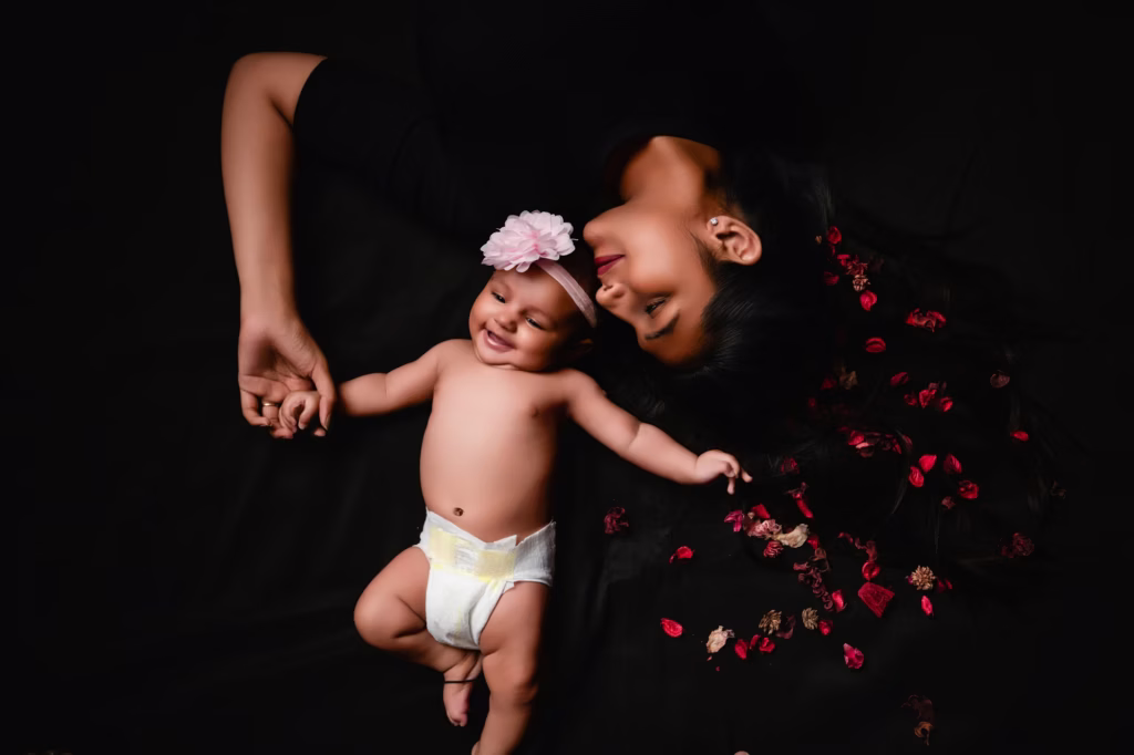 High-angle studio shot of mother and baby lying on black background baby in diaper with pink flower headband red flower petals scattered across mother's long dark hair