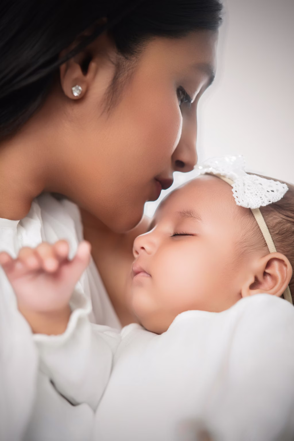 Mother kissing sleeping infant forehead white clothing lace headband soft lighting peaceful intimate tender moment studio