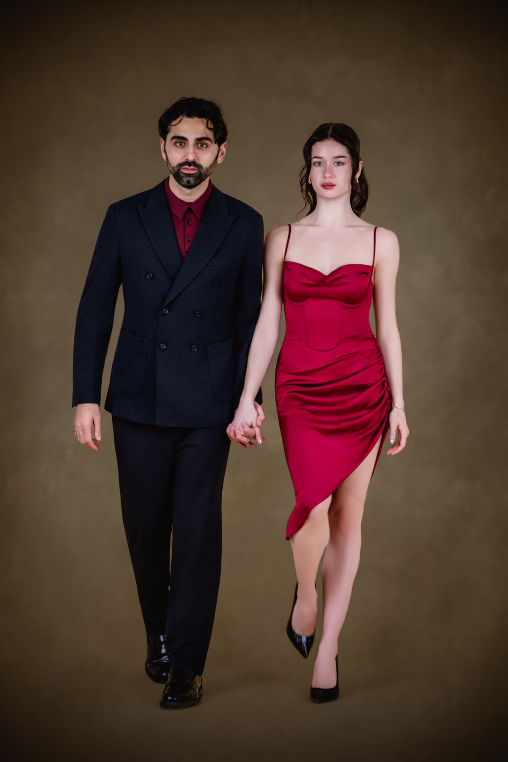 Formal full length studio portrait of couple standing hand in hand, man in black double breasted suit with burgundy shirt and woman in red satin cocktail dress with thigh high slit and black heels - beautiful couple photoshoot