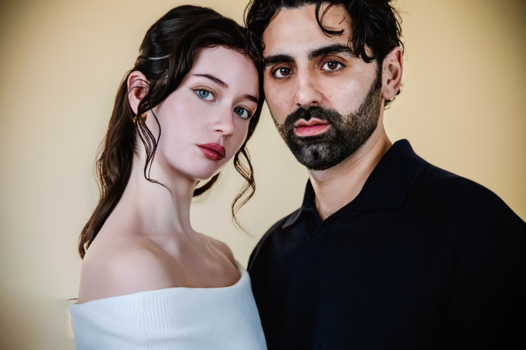 Tight headshot style portrait of couple leaning heads together, woman in off shoulder white top and man in dark polo shirt with soft even lighting against warm cream colored background - couples photoshoot