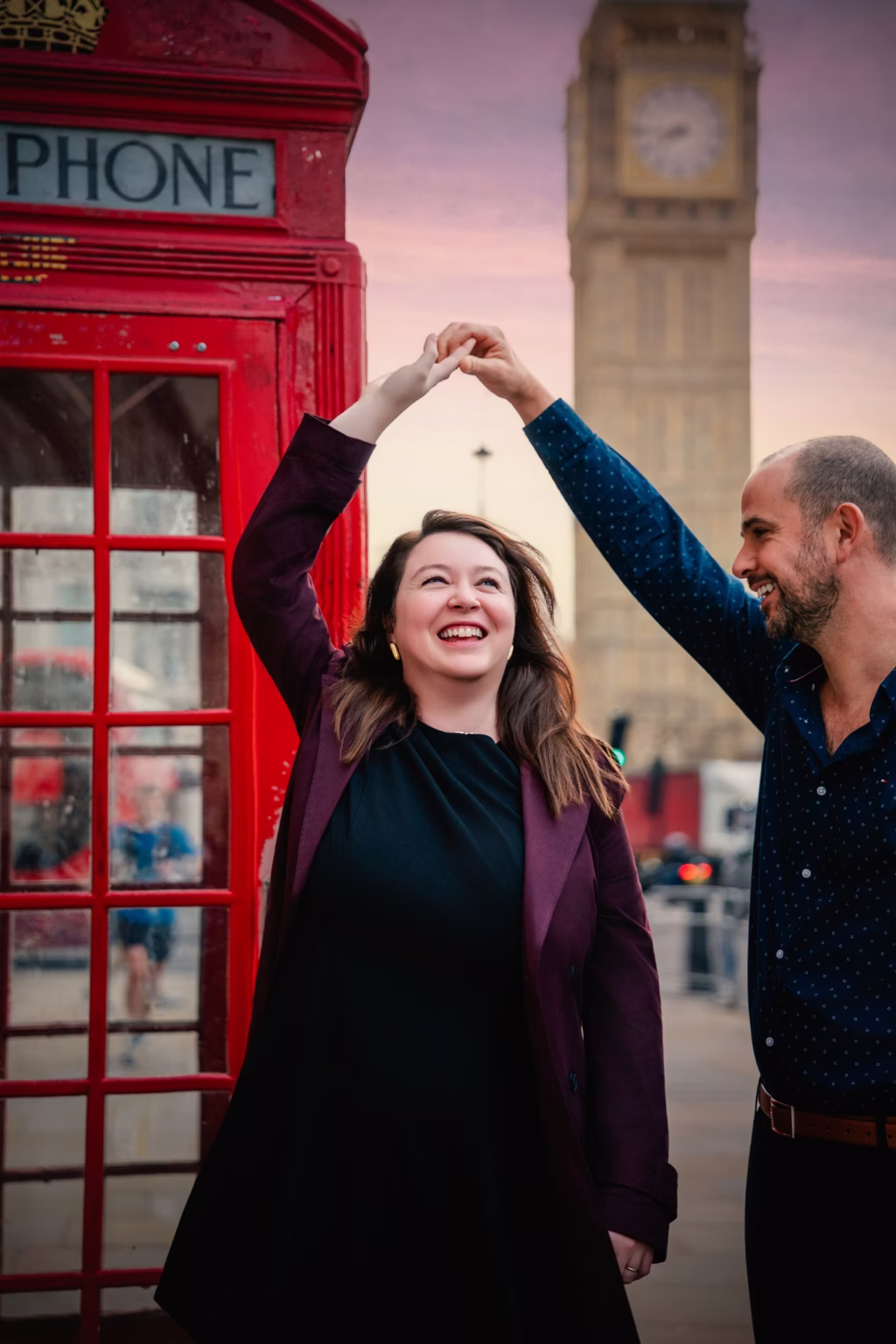 Candid outdoor shot of couple near iconic red London telephone box, man twirling woman who is laughing brightly with Big Ben visible in background under soft purple and pink sunset - couple photographers