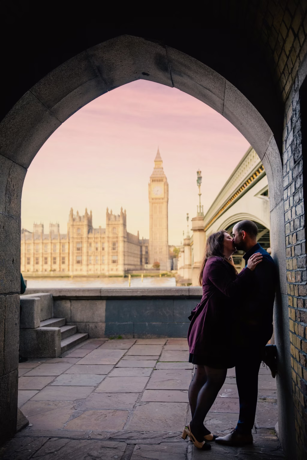 Romantic wide shot framed by stone archway on Thames embankment, couple sharing kiss with woman leaning against man, Houses of Parliament and Big Ben perfectly centered in background under twilight sky - couple photoshoot photographer