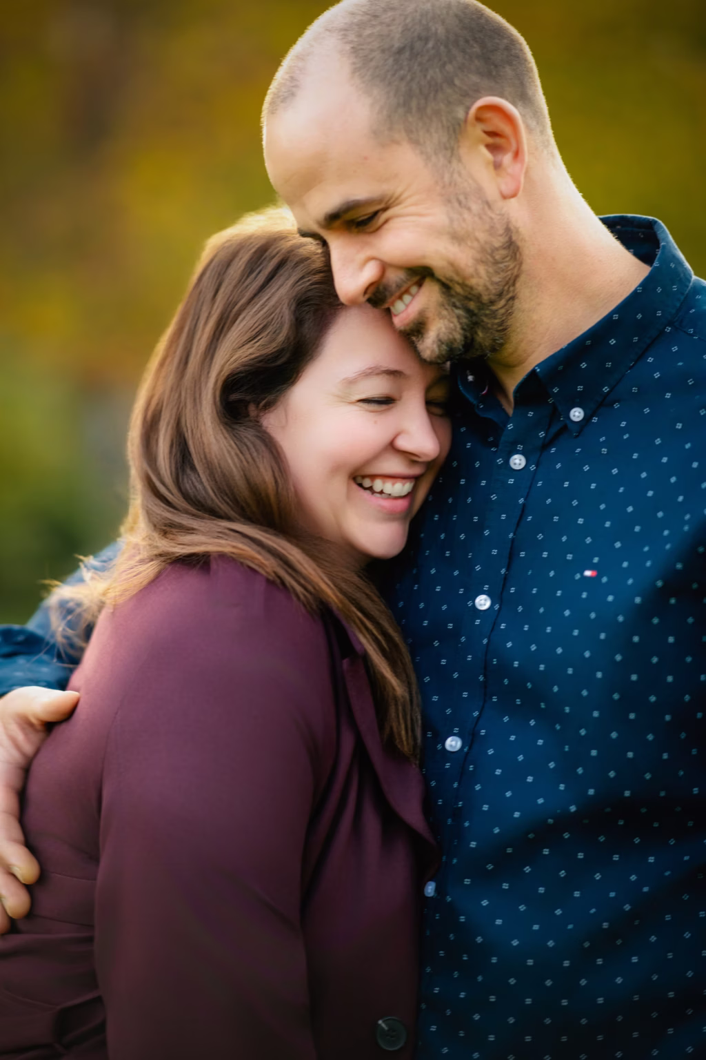 london couple photoshoots intimate outdoor close up of couple, man resting head against woman's with both eyes closed and wide happy smiles, soft warm bokeh of autumn foliage background