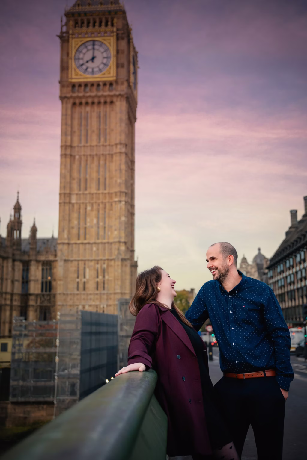 Candid outdoor shot of couple on bridge, woman leaning back and laughing while man looks at her with smile, iconic Big Ben Elizabeth Tower behind them under soft purple and orange twilight sky - couples photo session