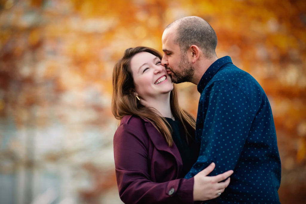 Man kissing woman on the cheek as she laughs, warm autumn bokeh behind them - couples photographer London