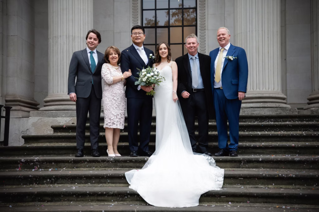 london elopement photographer formal group portrait of wedding party on stone steps with classical columns, bride in white lace gown holding green and white bouquet with groom in navy suit