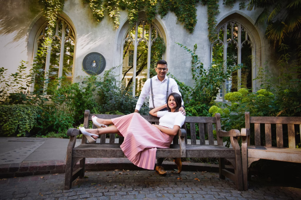 Lifestyle shot at St Dunstan in the East with woman reclining on wooden park bench legs up while man stands behind with hand on her shoulder against background of iconic ruined Gothic windows covered in climbing ivy - engagement photographer london