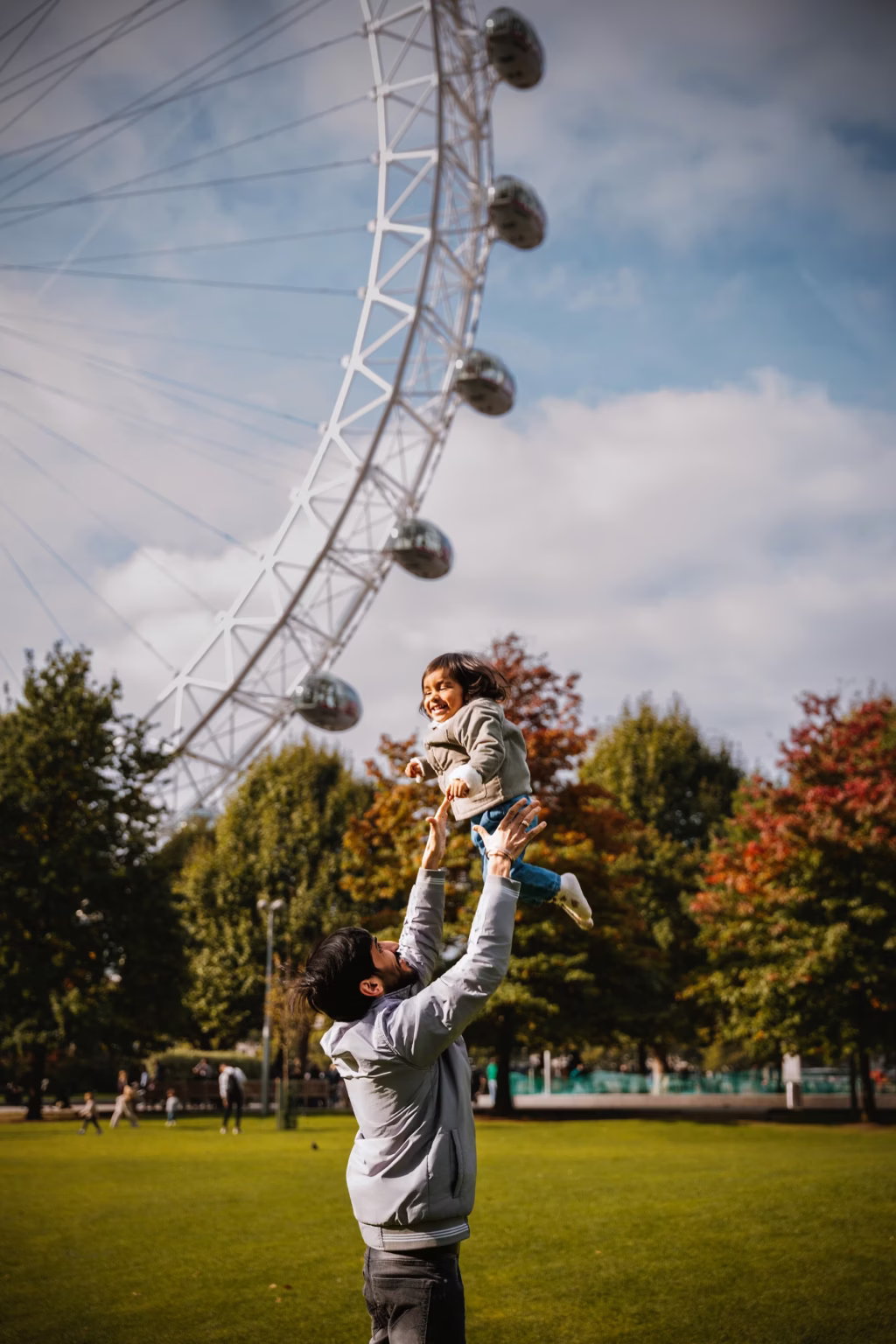 Father tossing young daughter in air park London Eye background blue sky white clouds autumnal trees grassy area action joy