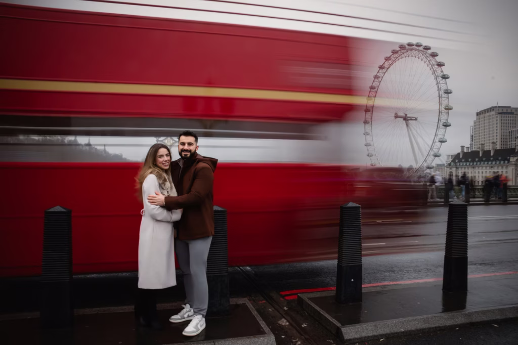 Long-exposure shot of couple standing on London sidewalk red double-decker bus blurred into streak of motion behind them London Eye visible in distance