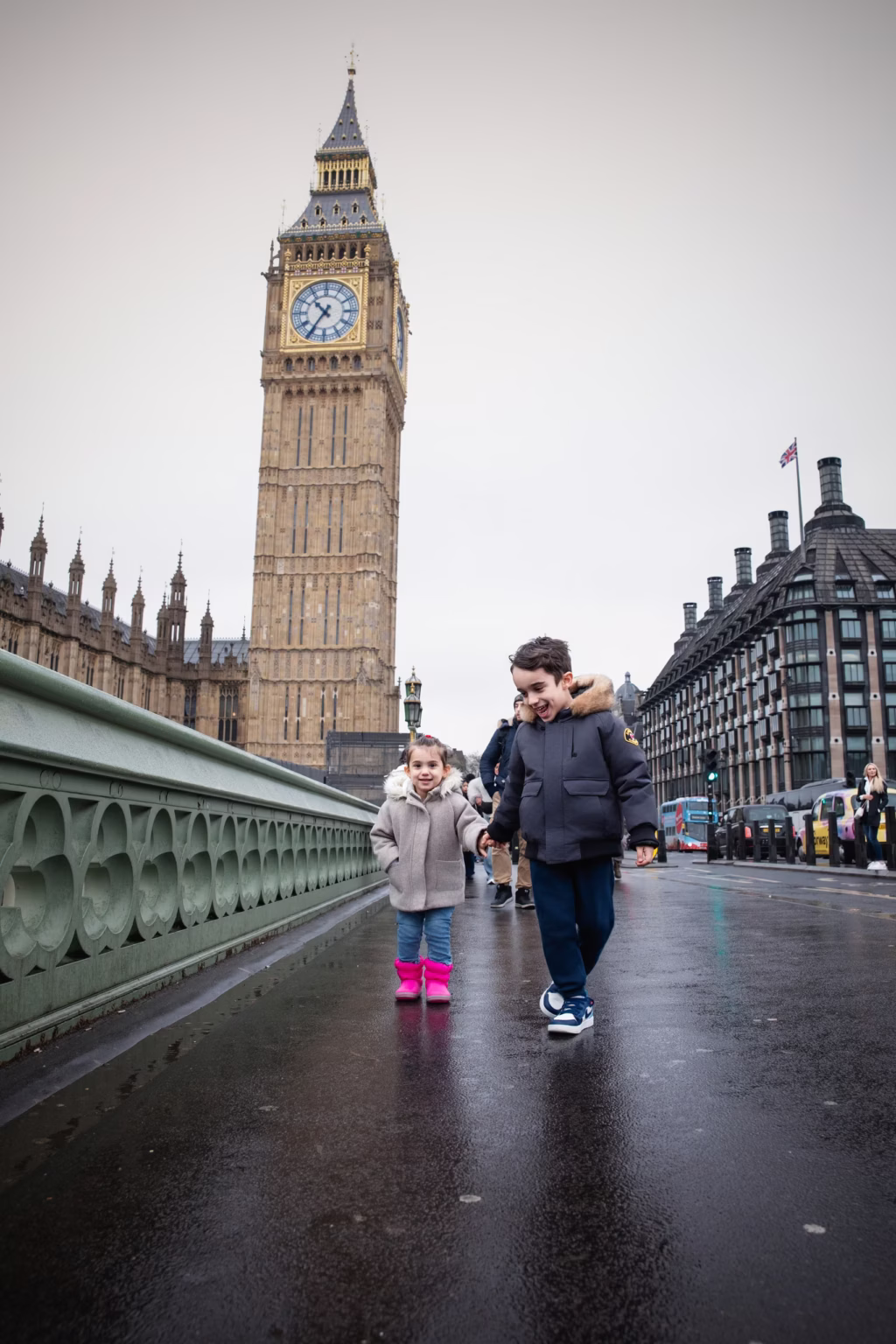 Two young children boy and girl holding hands walking along Westminster Bridge boy in dark parka girl in beige coat and pink boots Big Ben in background - family photoshoot london