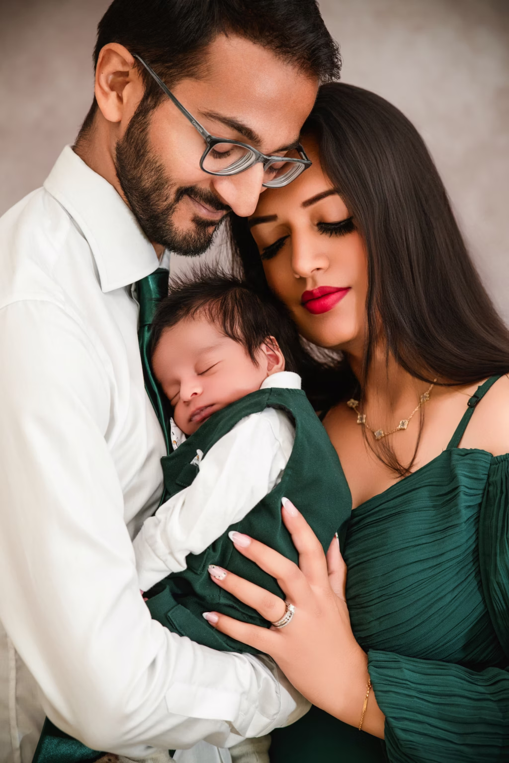 Intimate studio close-up of father and mother holding sleeping newborn father wears green tie mother has red lipstick baby swaddled in dark green - family photographer london