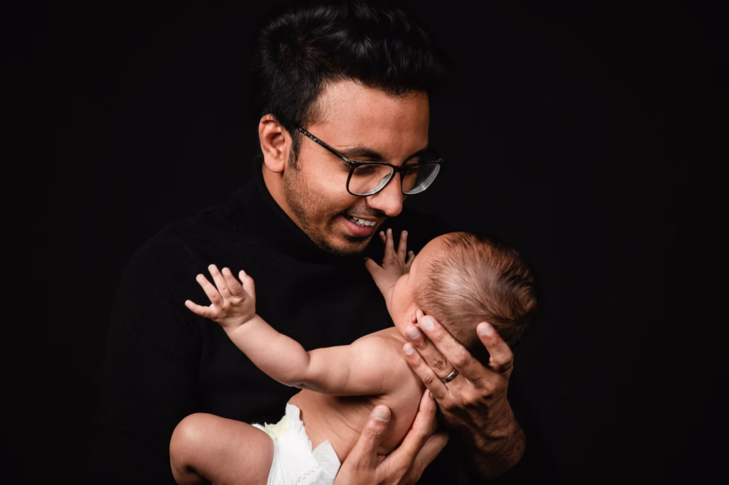 High-contrast studio portrait against black background father wearing glasses and black turtleneck smiling down at baby he is holding in his hands - family portrait photography