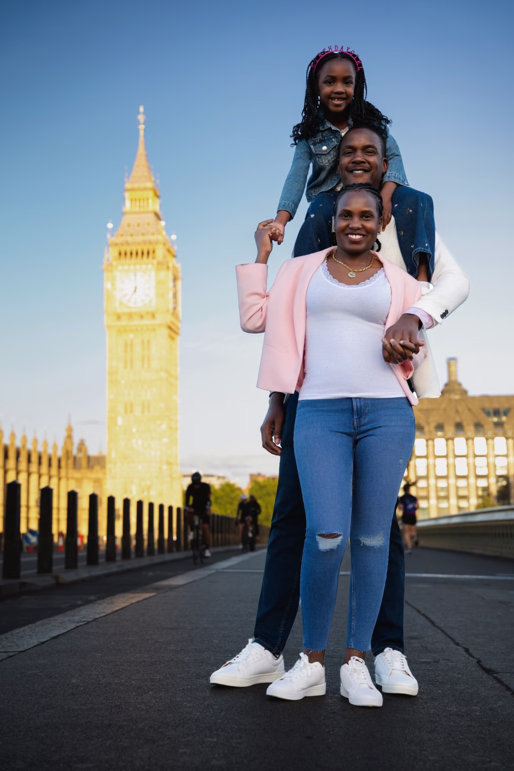 Full-length family portrait on Westminster Bridge father has young daughter on shoulders standing next to mother Big Ben visible and illuminated in background - family photoshoot london
