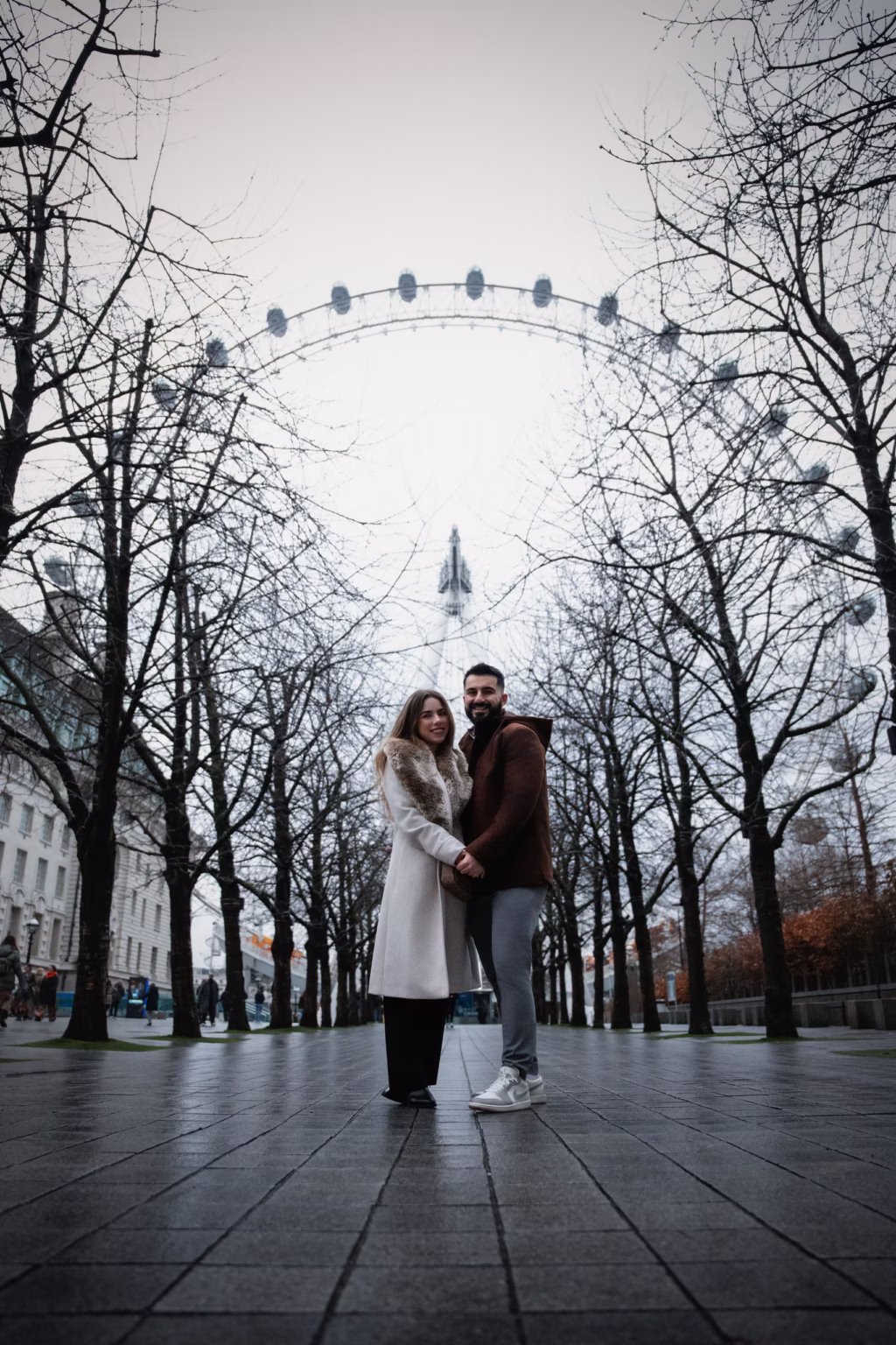 Couple standing in center of tree-lined path near South Bank London Eye centered in background behind bare winter trees