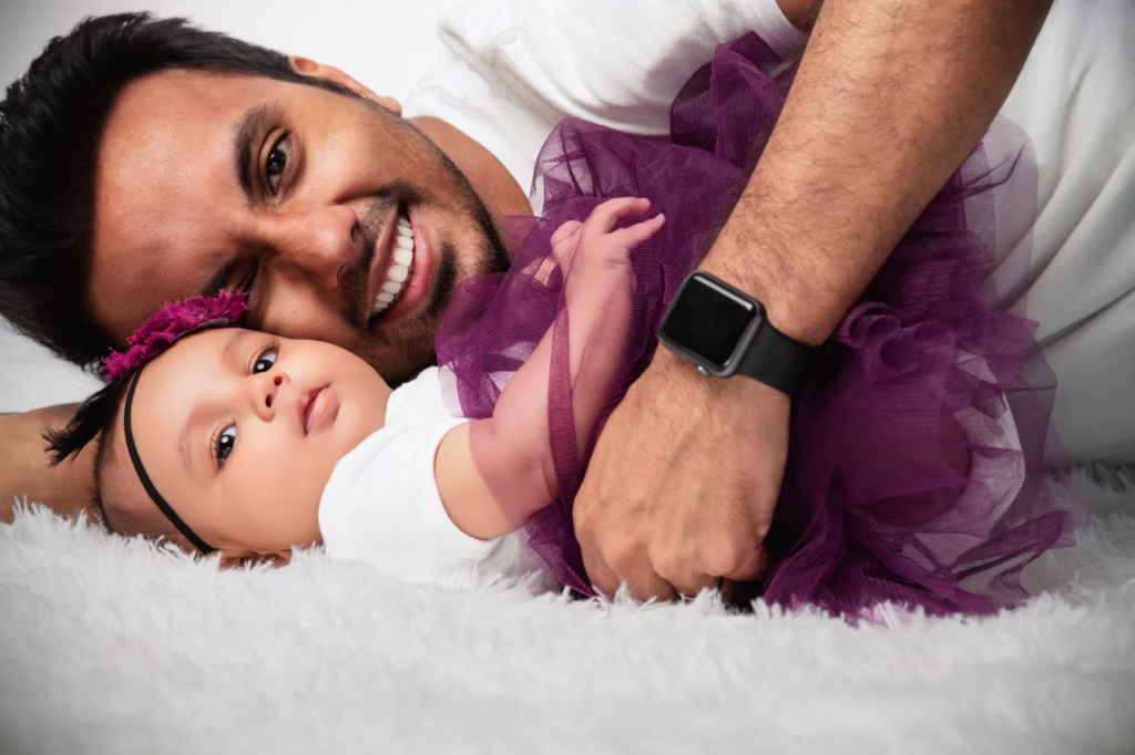 Close-up high-key shot of father and baby girl on fluffy white rug, father smiling broadly at camera, baby in purple tulle tutu with floral headband looking toward lens with curious expression - family portrait studio, father baby playful