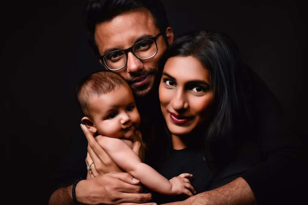 Dramatic high-contrast studio portrait of mother and father holding baby against solid black background, father wears glasses, family posed closely together creating moody intimate feel - family photography studio, black background moody