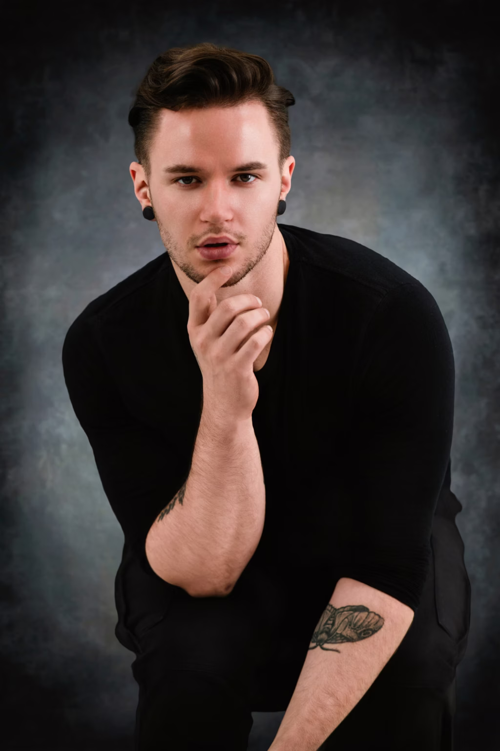 London Fashion Photographer Studio - Centered studio portrait man styled brown hair black gauge earrings black crewneck top one hand chin mottled dark charcoal grey background