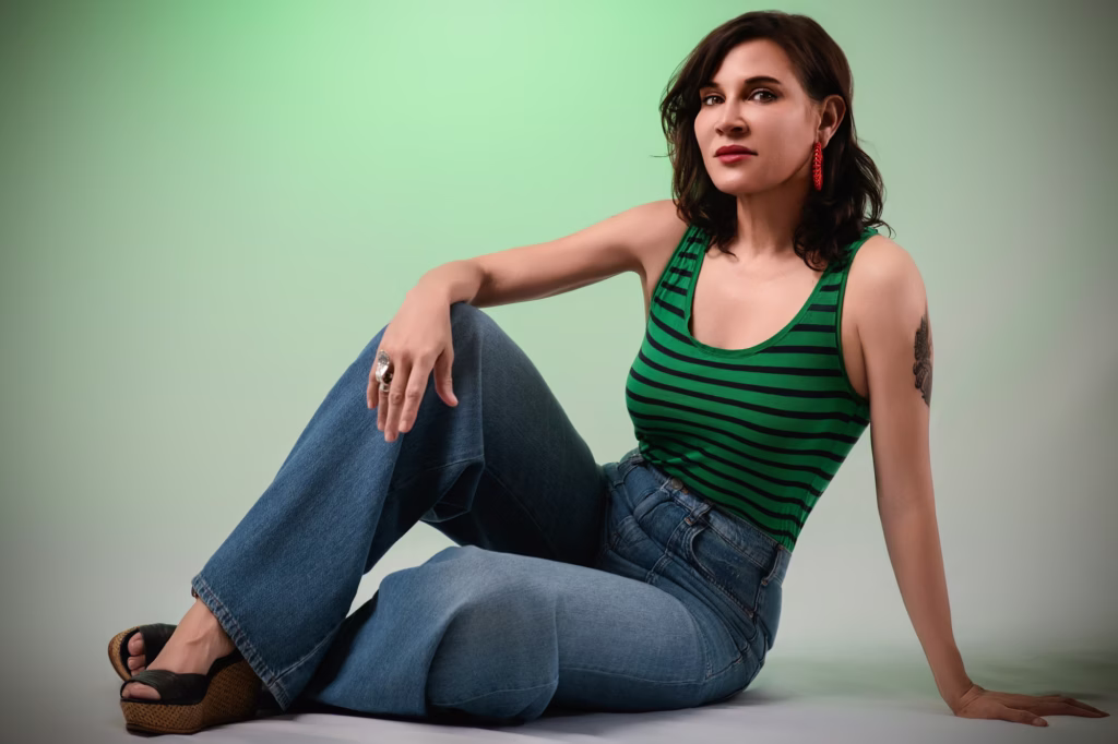 London Fashion Photography London - Full-length studio pose woman sitting floor soft green gradient background green black striped tank top wide-leg blue jeans platform wedge sandals large silver ring red statement earring