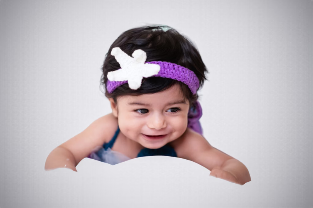 Bright studio portrait of smiling infant girl in purple crocheted headband with white starfish accent, leaning over white cloud-shaped prop against soft grey background - london infant photography starfish headband smile