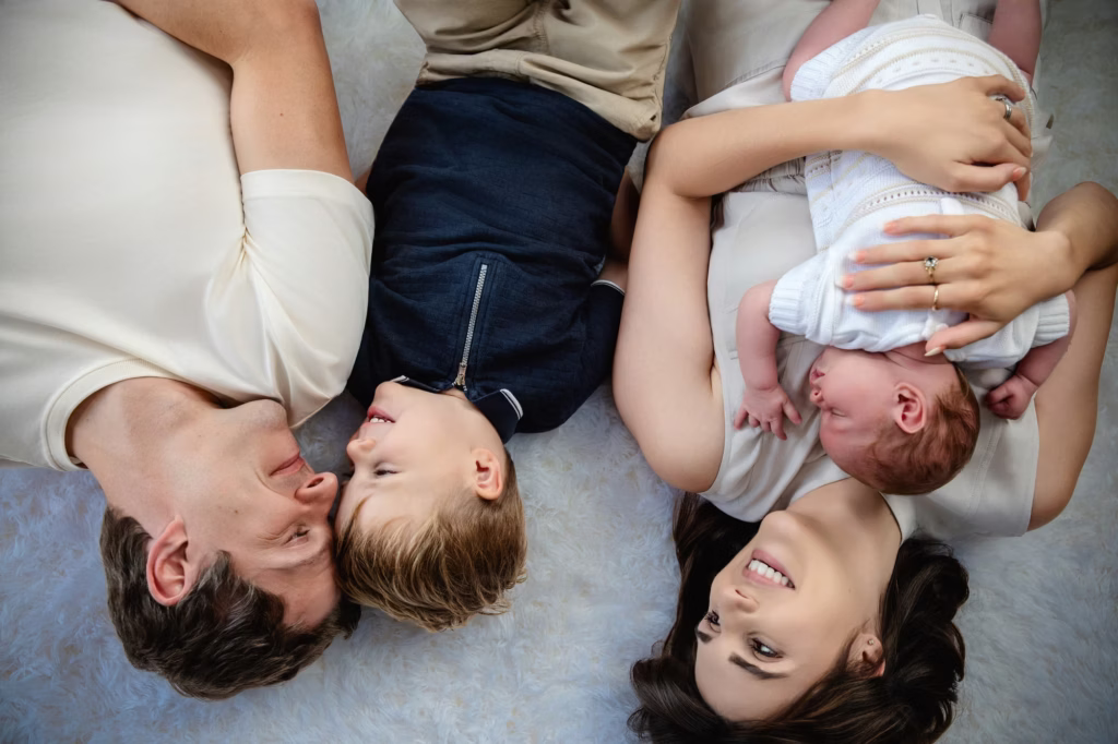 Overhead lay flat family portrait on white shag rug, father older son mother and sleeping newborn side-by-side, mother cradling newborn in white knit outfit - new born photo shoots, infant photography family lay flat