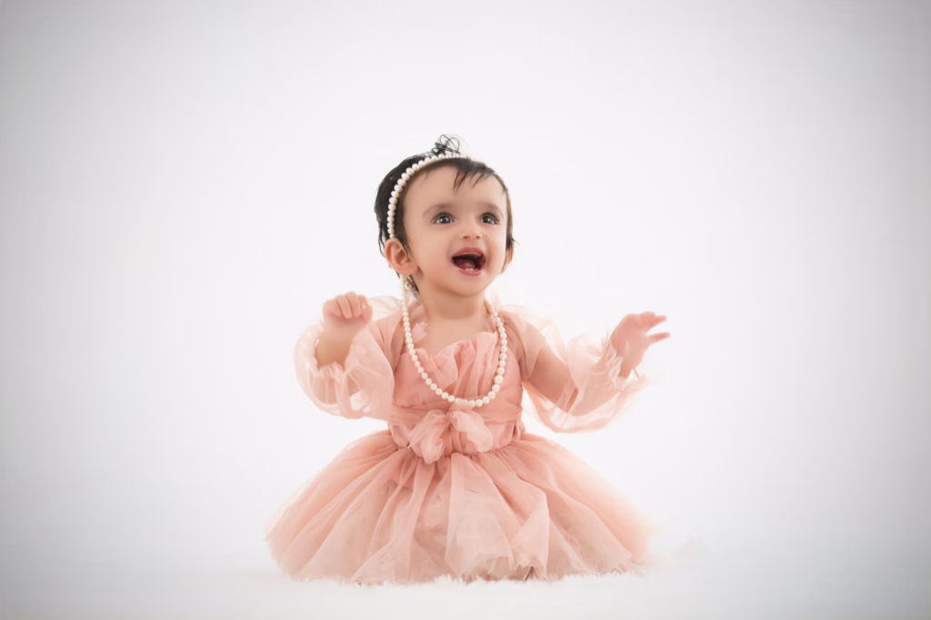 High-key studio portrait of infant girl on white furry surface, peach-colored voluminous tulle dress, pearl headband with matching long pearl necklace, open-mouthed laugh - london infant photography peach tulle pearl