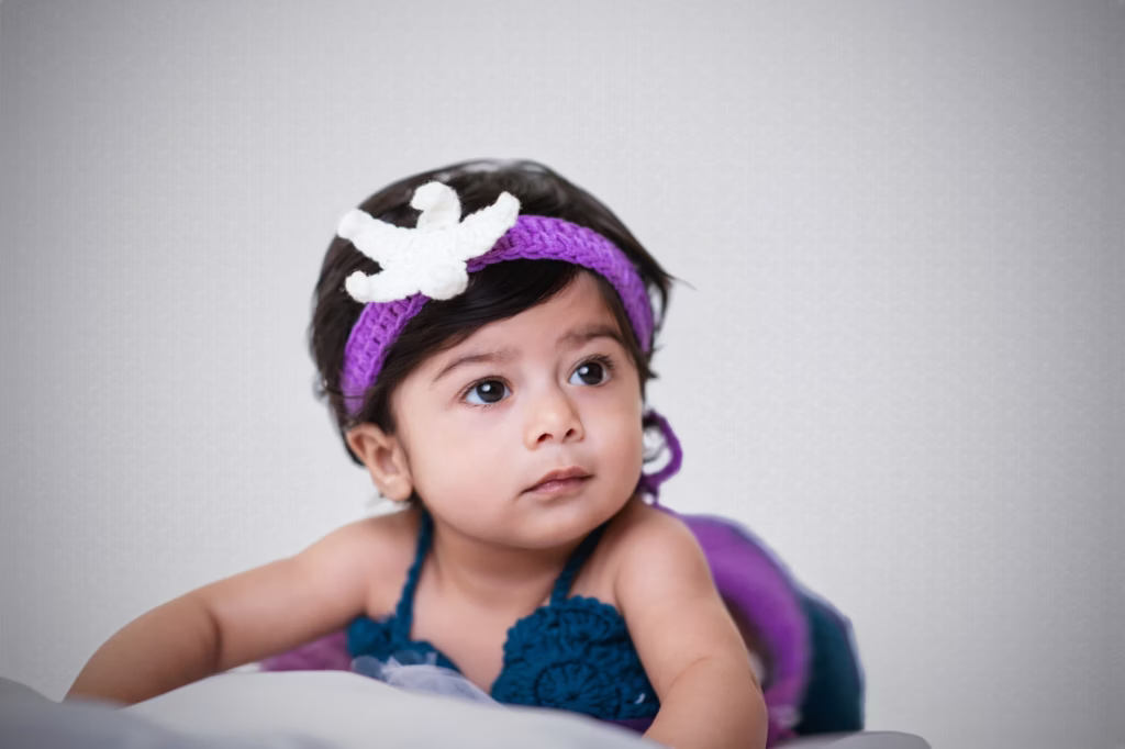 Close-up studio portrait of infant girl in purple starfish headband, looking off to side with curious and serene expression, resting arms on soft white surface - london infant photography starfish close-up serene