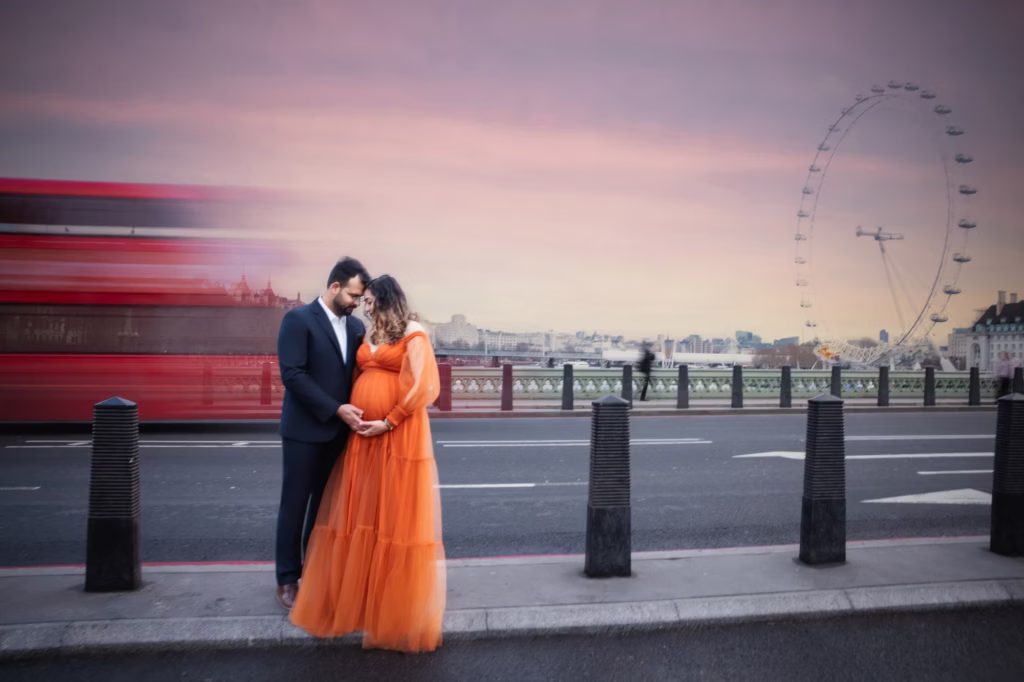 Couple on Westminster Bridge at dusk, woman in flowing bright orange tulle gown, red double-decker bus in motion, London Eye visible against pink sky - london maternity urban portrait