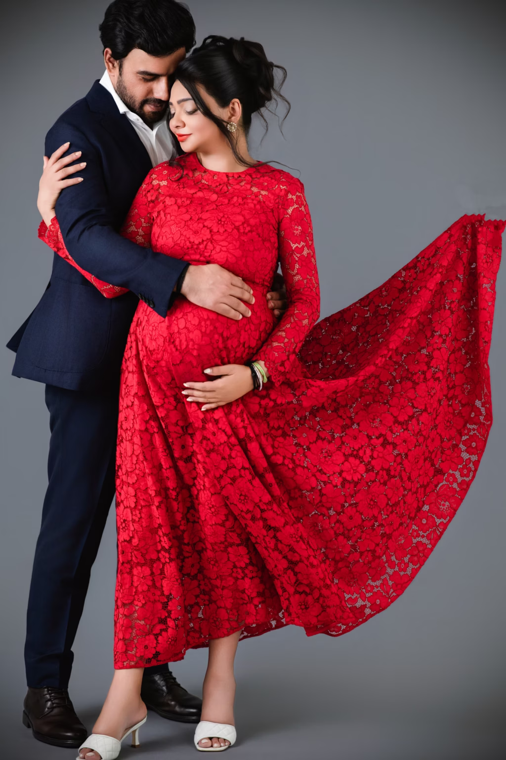 Studio shot against solid grey background, man in navy suit embracing woman in vibrant red lace gown, long skirt of her dress captured mid-air billowing out to side for dramatic effect - studio maternity photography, studioshoot dramatic flowing dress embrace