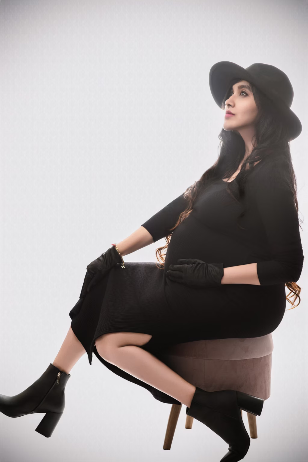 High-fashion studio portrait, woman in black long-sleeved dress, black gloves, black wide-brimmed hat sitting on velvet stool, posed in profile against bright white background looking upward with serene expression - studio maternity photos, studioshoot high fashion gloves hat