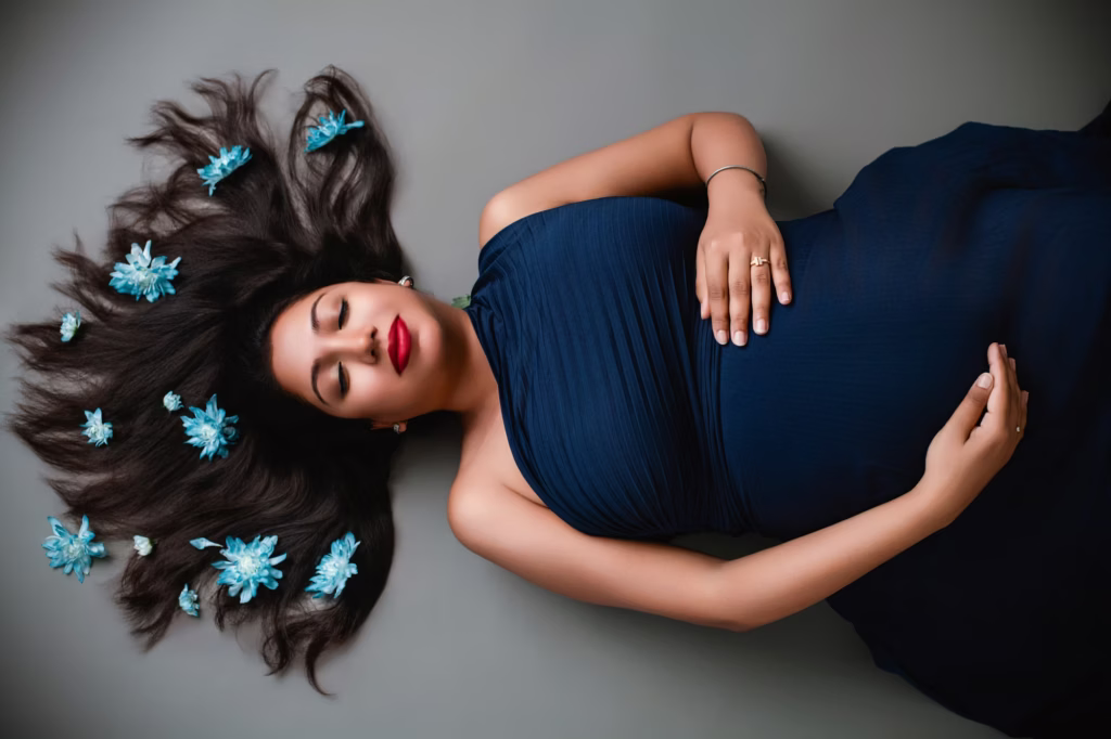 Top-down artistic shot of woman lying on grey surface with long dark hair fanned out adorned with light blue flowers, eyes closed, hands resting on pregnant bump - london maternity photography creative pose
