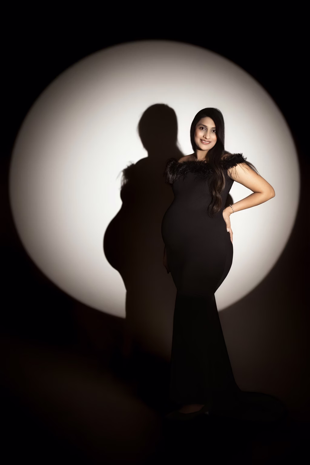 Circular spotlight portrait with woman standing facing forward in black feathered dress, one hand on hip other on belly, silhouette projected on illuminated background - maternity photography london, standing pose dramatic lighting