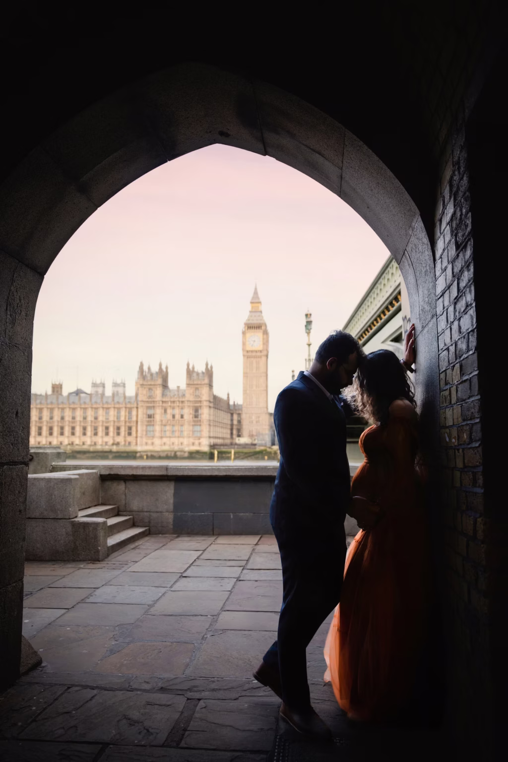 Cinematic outdoor shot from within stone archway near Thames, couple silhouetted in foreground facing each other intimately, Houses of Parliament and Big Ben perfectly framed through arch against soft pink sky - maternity photographer, architectural framing