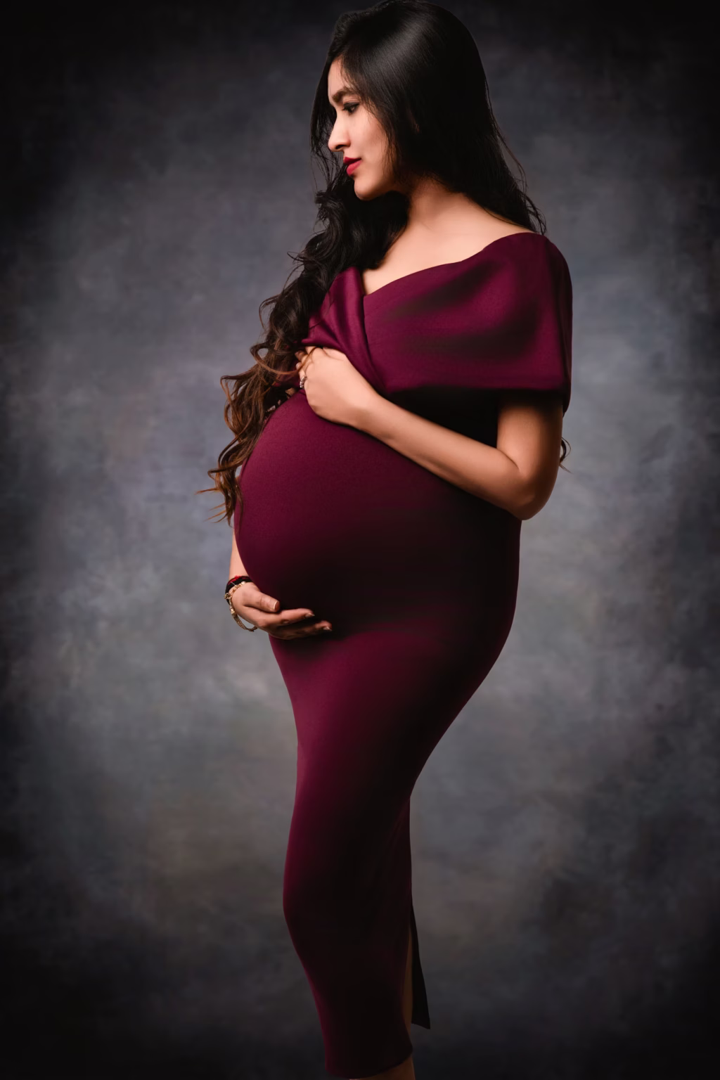 Sophisticated studio portrait of woman in profile against textured dark grey background wearing form-fitting off-shoulder gown in deep plum/burgundy color, cradling bump with both hands - photographers maternity, elegant profile