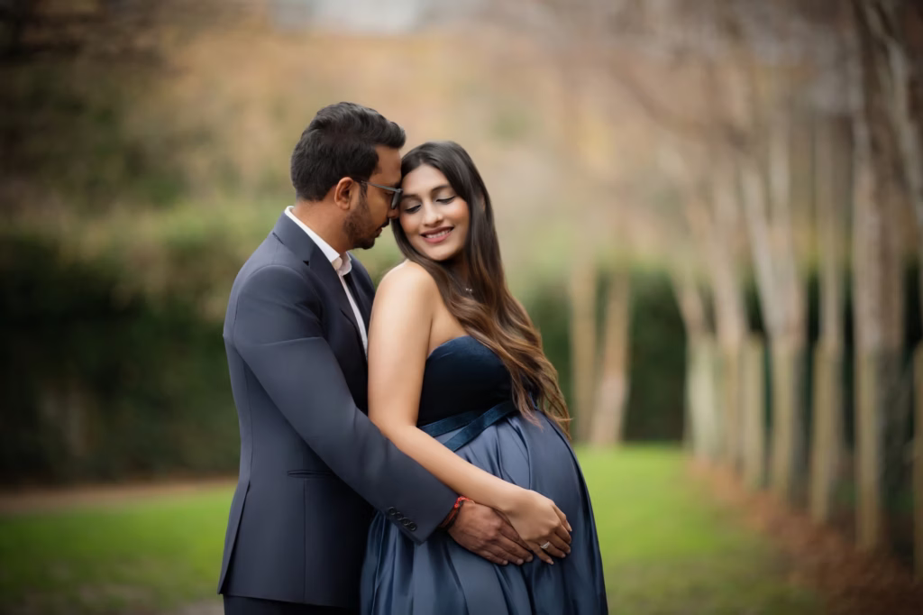 Intimate outdoor portrait in park with blurred trees, man in suit standing behind woman in strapless navy velvet gown leaning head against hers, both resting hands on pregnant belly - photographer maternity, park intimate