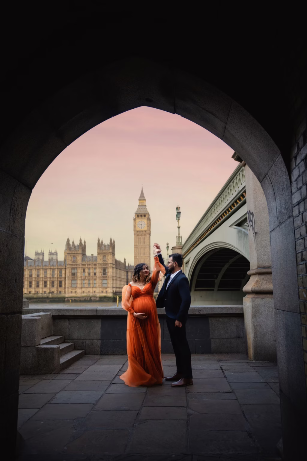 Romantic shot framed by stone archway along South Bank, couple dancing intimately, woman in vibrant orange tulle gown, man in dark suit, Big Ben and Houses of Parliament perfectly centered in background under pink sky - maternity photographer london, archway dance South Bank